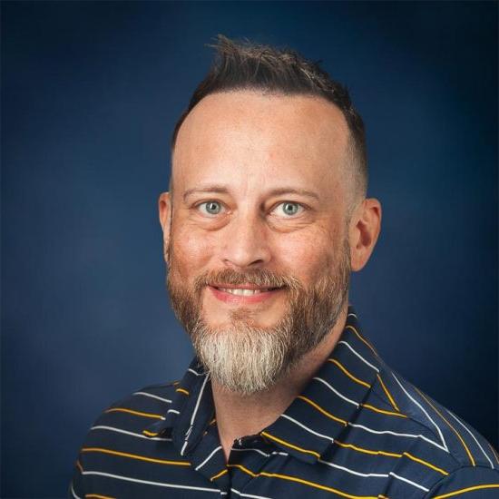 Portrait of a smiling man with a short dark hair and a beard, wearing a blue with colorful strips, is set against a dark blue background.